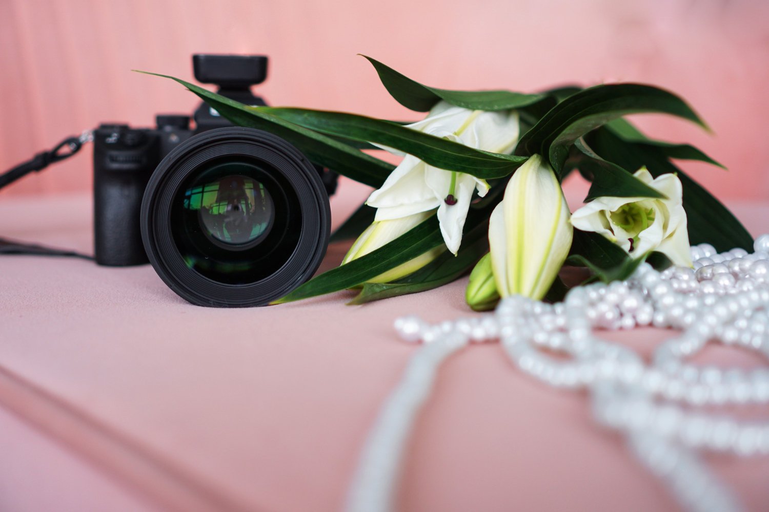 Second Photographer & Videographer Add-Ons: What They Actually Cost and Cover 11 Close-up of a camera lens beside white lilies on a pink backdrop, representing a second wedding photographer