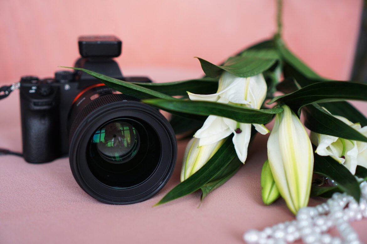 Professional camera with white lilies and a pearl necklace on a soft pink background, symbolizing wedding photography add-ons