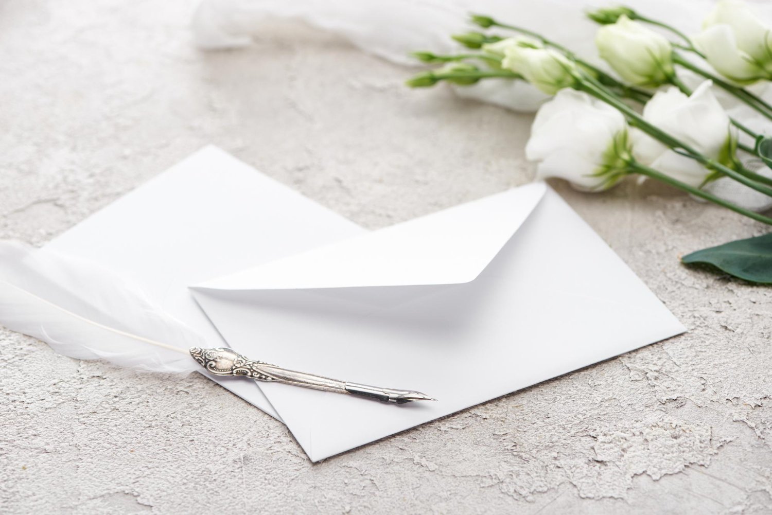 Plus-One Etiquette for Weddings: Who Gets Invited and Who Doesn't 13 White envelope beside eustoma flowers and a quill pen on a textured surface, representing RSVP card design