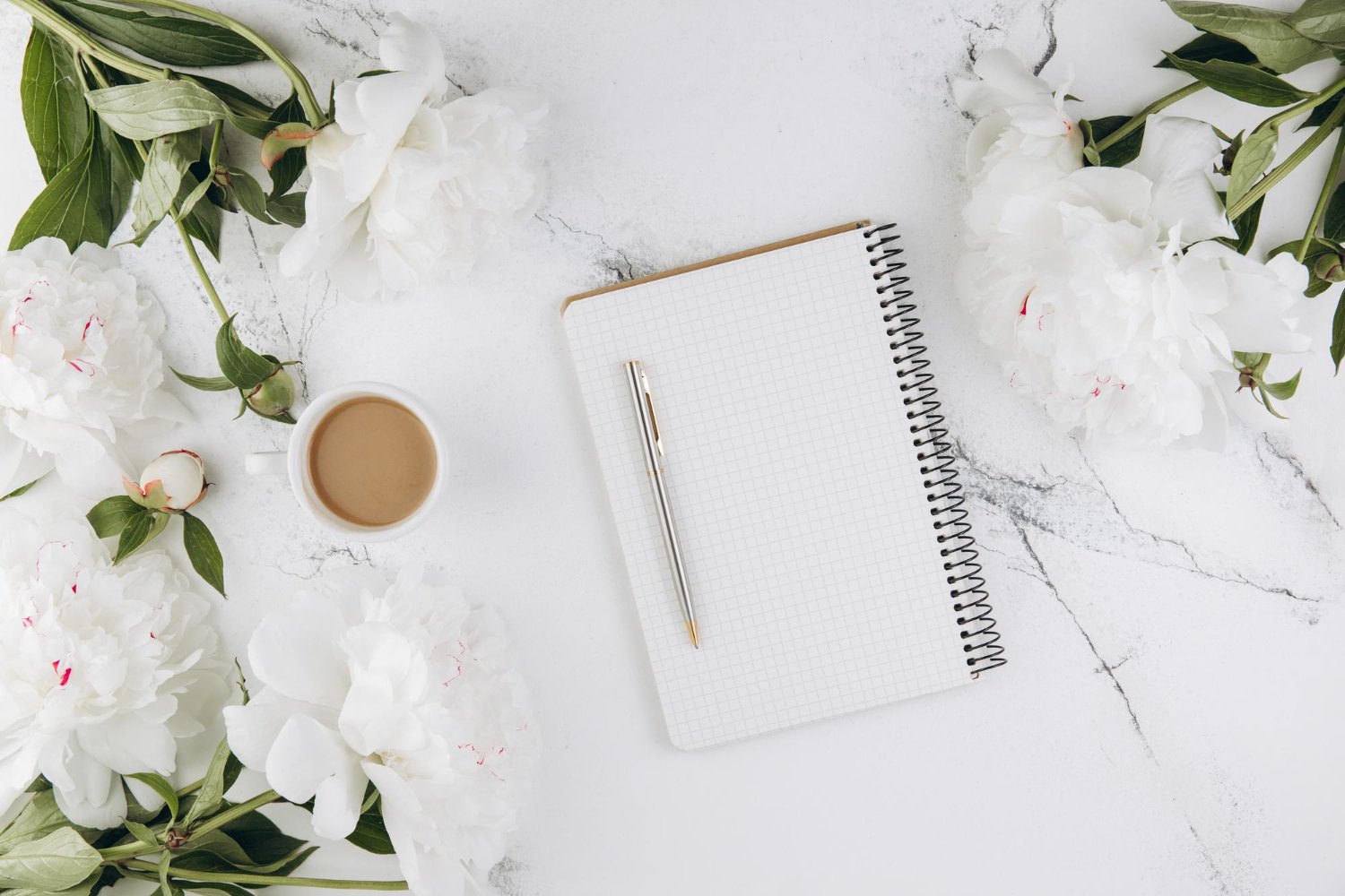 Plus-One Etiquette for Weddings: Who Gets Invited and Who Doesn't 12 White peony flowers beside an open notebook and pen on a marble surface for wedding guest list planning